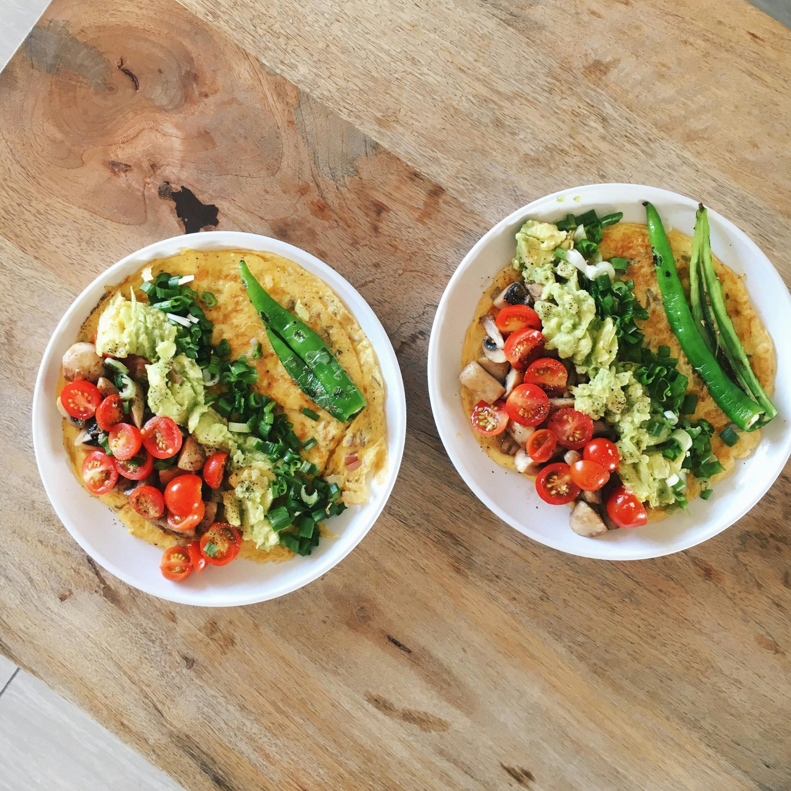 Two delicious omelette plates with avocado, tomatoes, and greens on a wooden table.